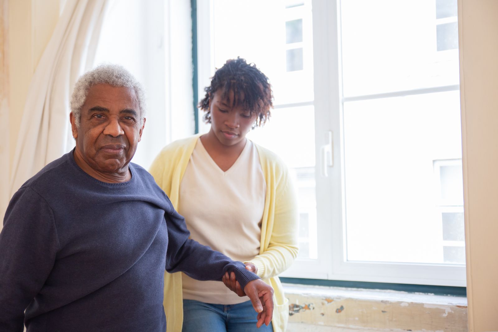 A carer supporting an older man beside a bright window.