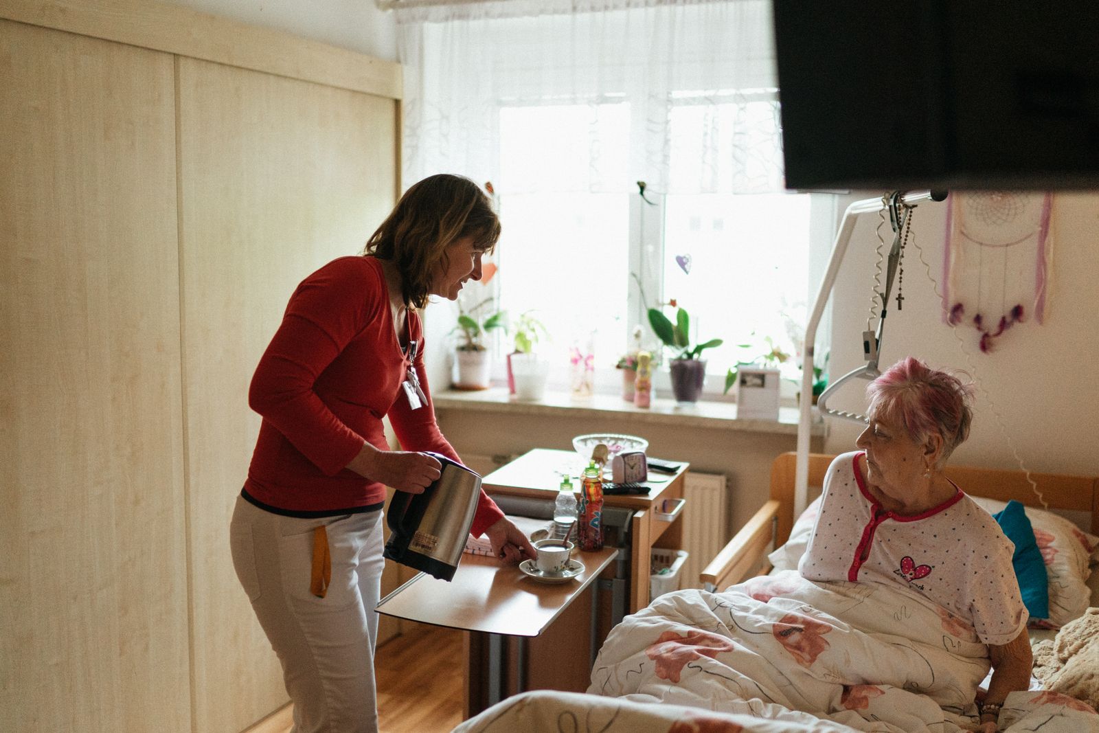 A carer bringing a warm drink to an older woman resting in her bedroom.